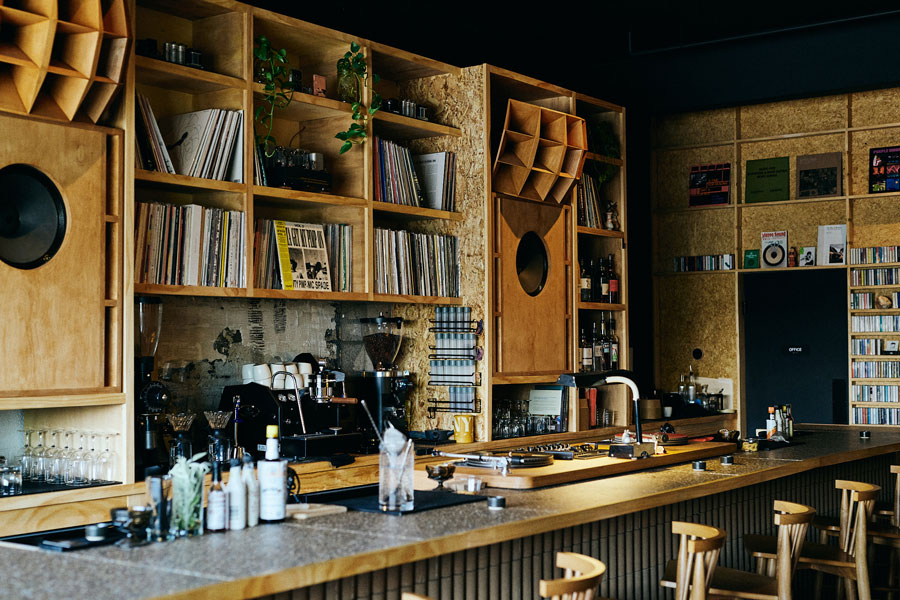 Behind-the-bar view of Mono Tape Club with wooden shelving full of vinyl and CDs, large round horn speakers, an espresso machine, and bar stools along the concrete counter.