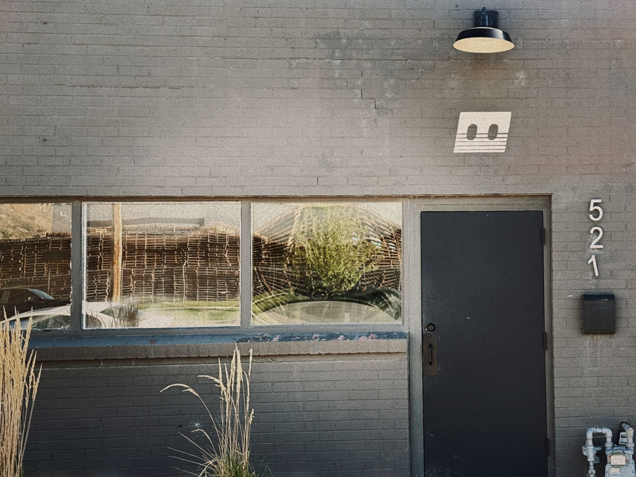 Gray brick exterior of Mono Tape Club at 521, with a dark entry door, large storefront window, gooseneck light, and the club's cassette-style logo mounted on the wall.