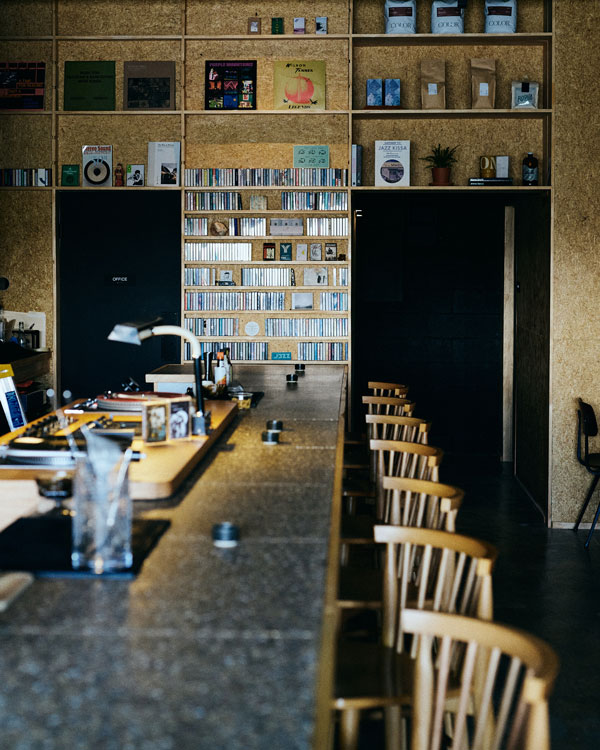 View down the Mono Tape Club bar toward a tall wall of shelving lined with cassettes and CDs, wooden chairs pulled up to the counter.